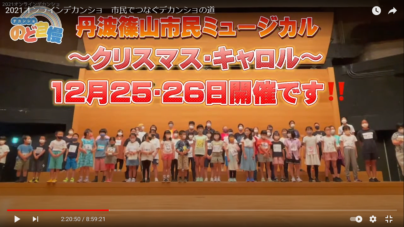 舞台上に市民ミュージカル出演者がそろっていて、上部にクリスマスキャロル開催のお知らせが書いてある。