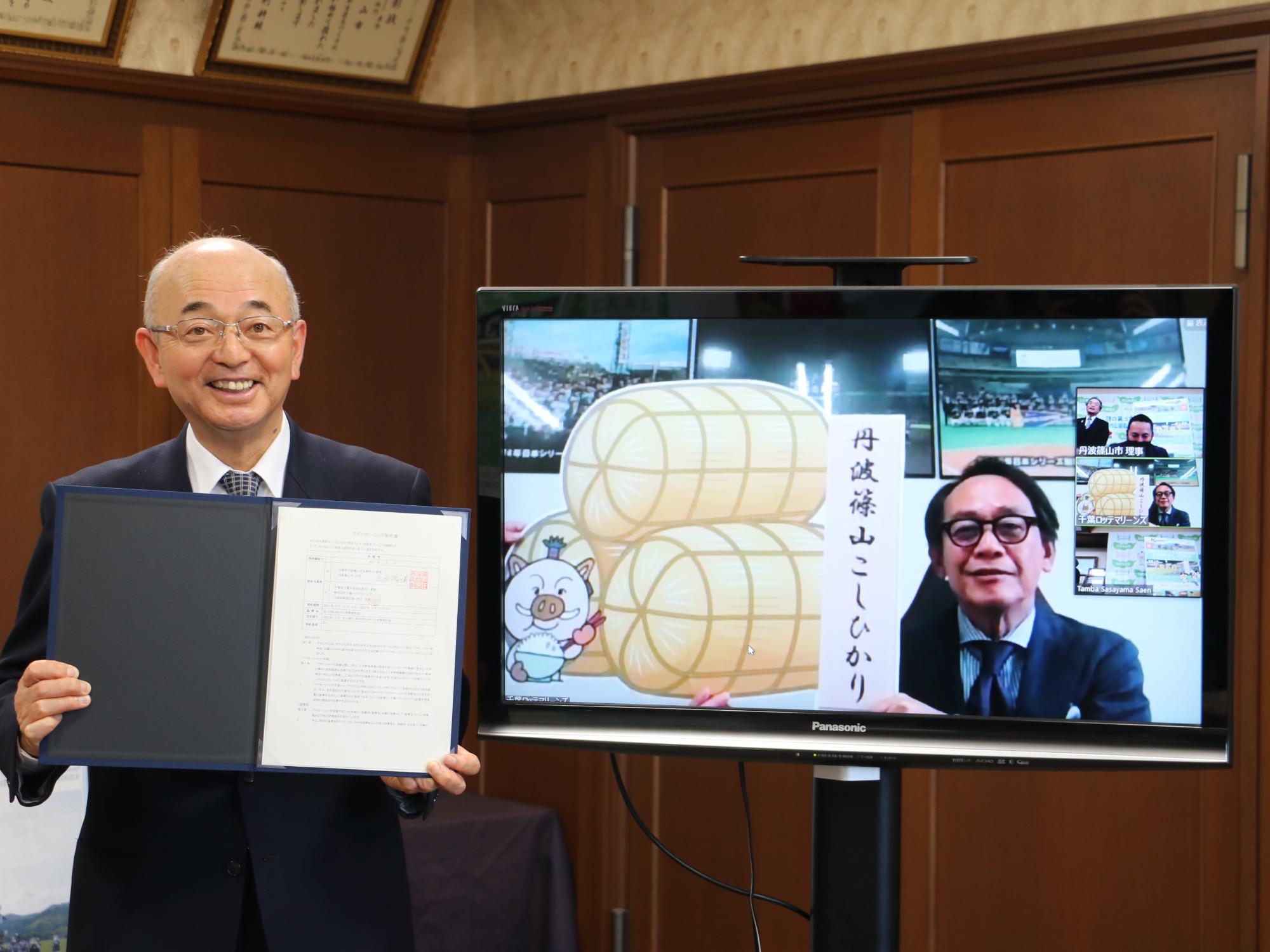 市長が署名・押印した契約書を手に持っている。モニターの河合社長はこしひかりのプレートを持っている。