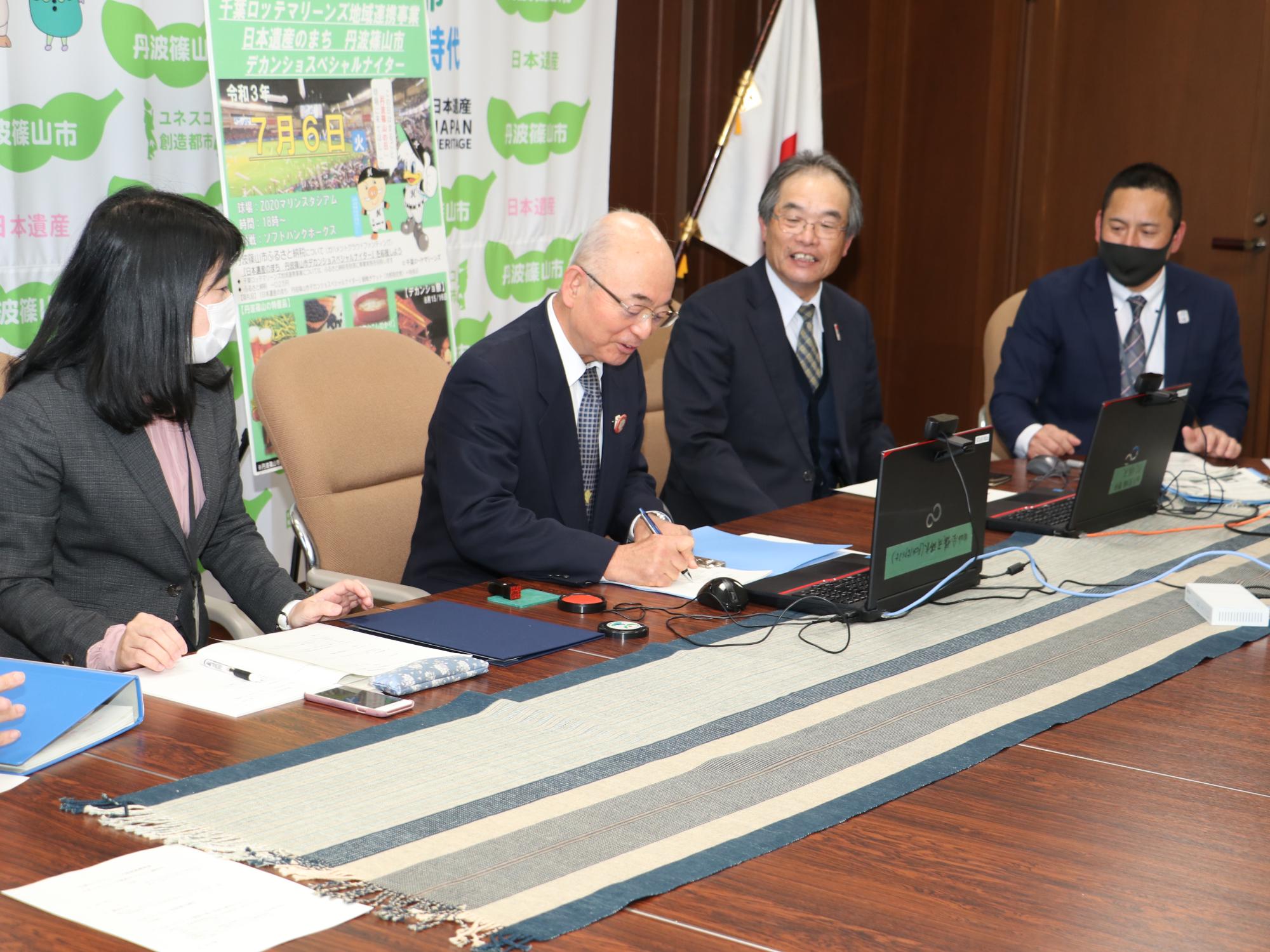 市長が契約書に署名している。周りの参加者が見守っている。