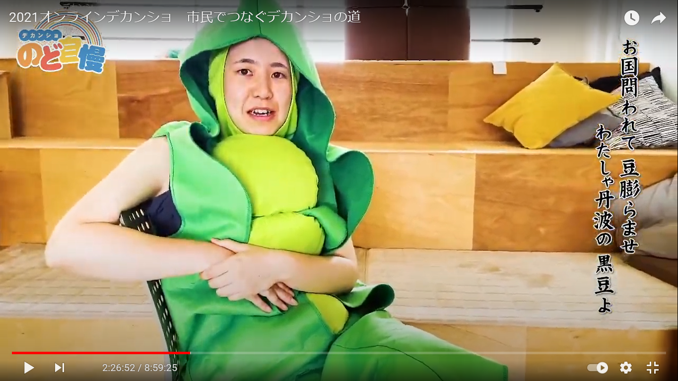 枝豆の着ぐるみを着た男性が椅子に座ってデカンショ節を歌っている
