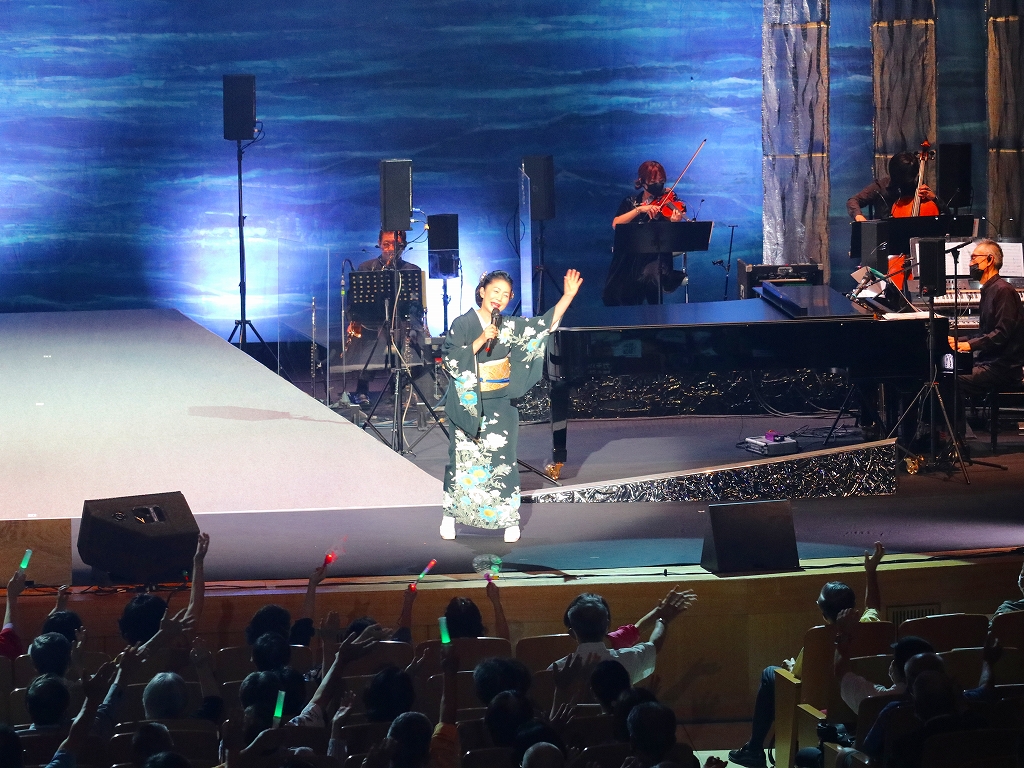 舞台に着物を着た女性がマイクを持って歌っている。後ろにはピアノや弦楽器奏者がいる。客席はペンライトを振っている。