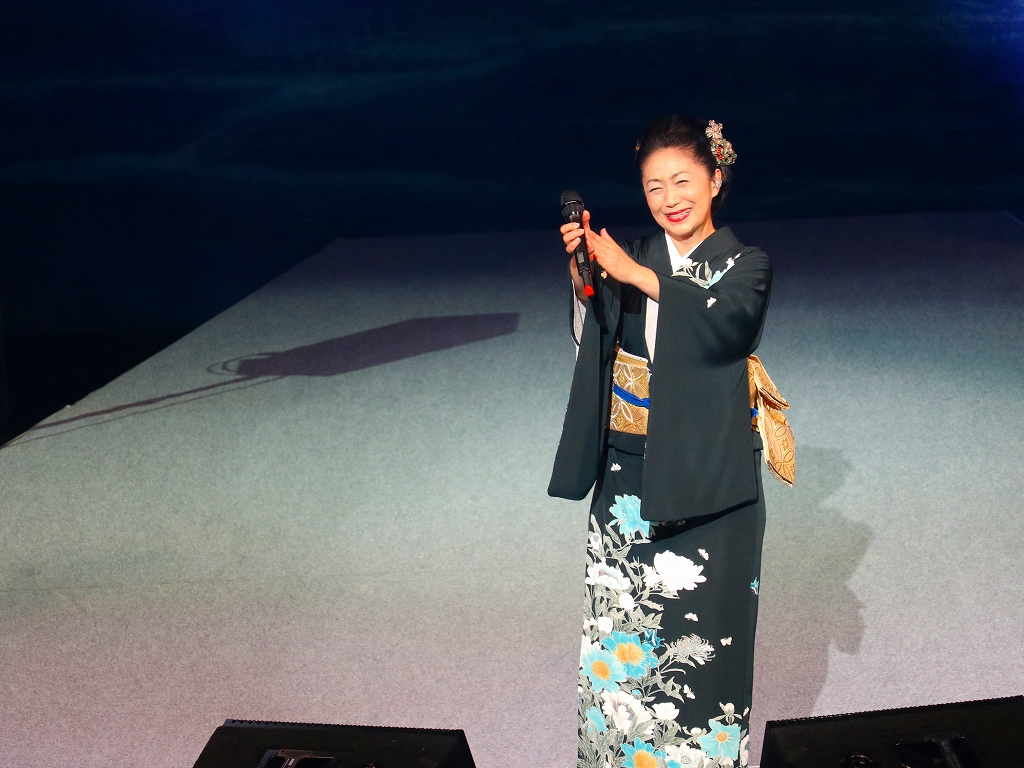 着物を着た女性がマイクを持ち客席に向かって拍手している。