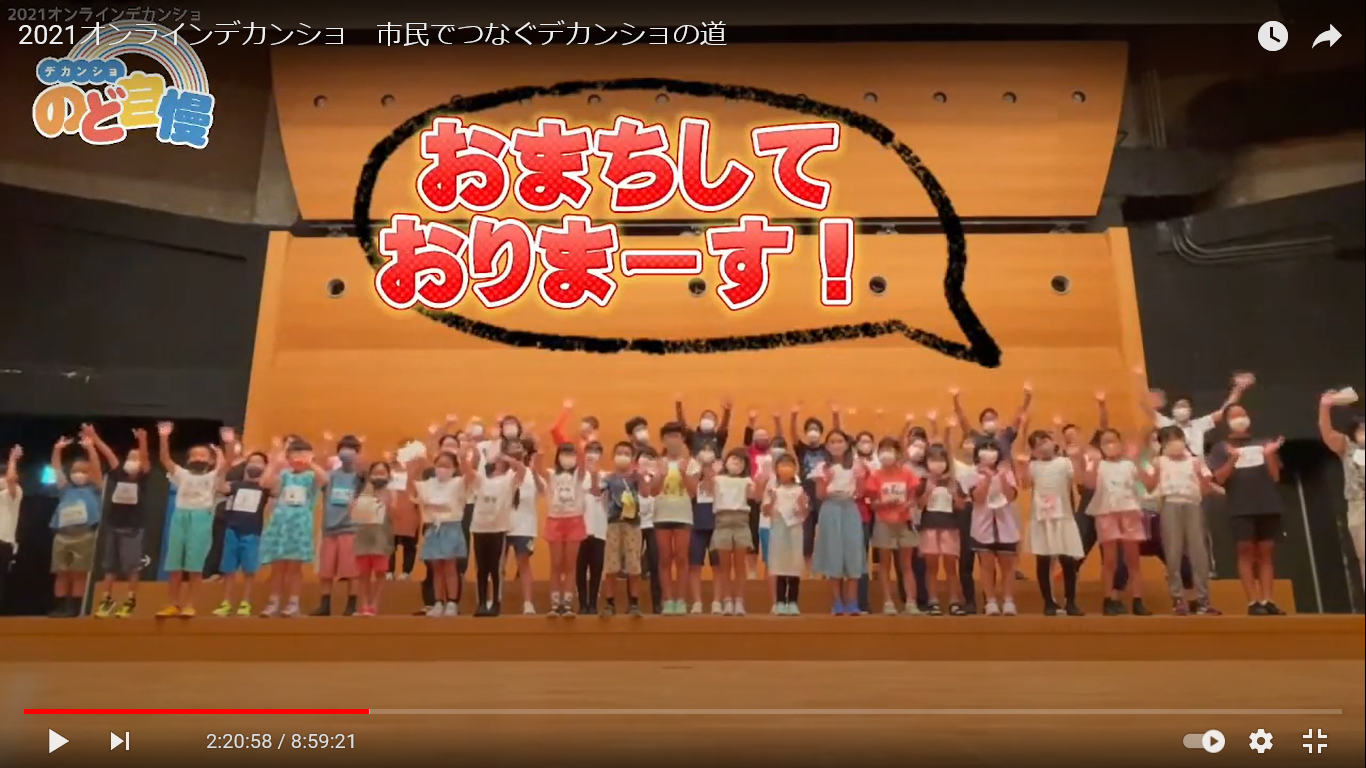 舞台上に市民ミュージカル出演者がそろって手を振っている。上部におまちしておりまーす！と書いてある。