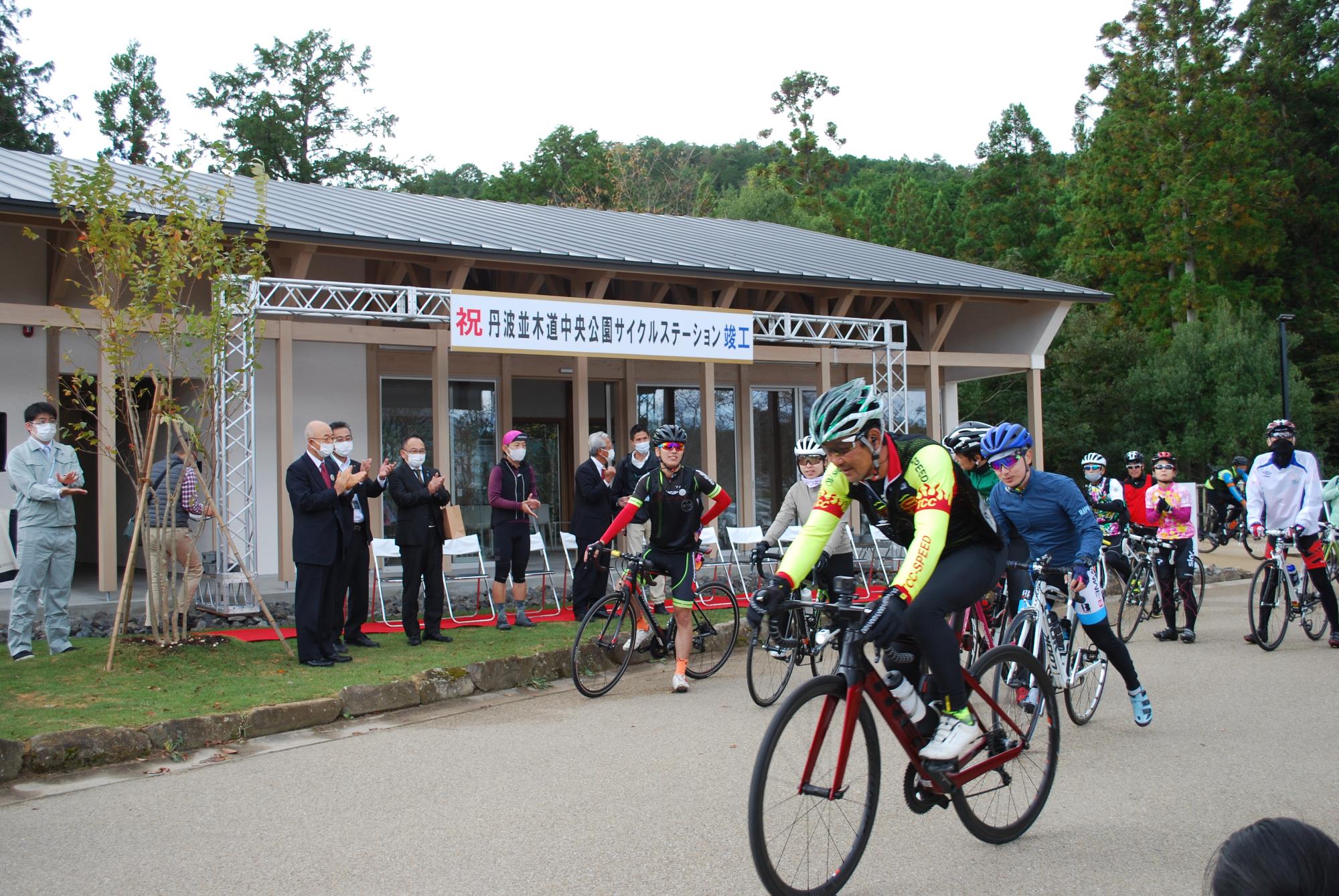 ヘルメットをかぶり自転車に乗ったレーサーたちがサイクルステーションを出発している
