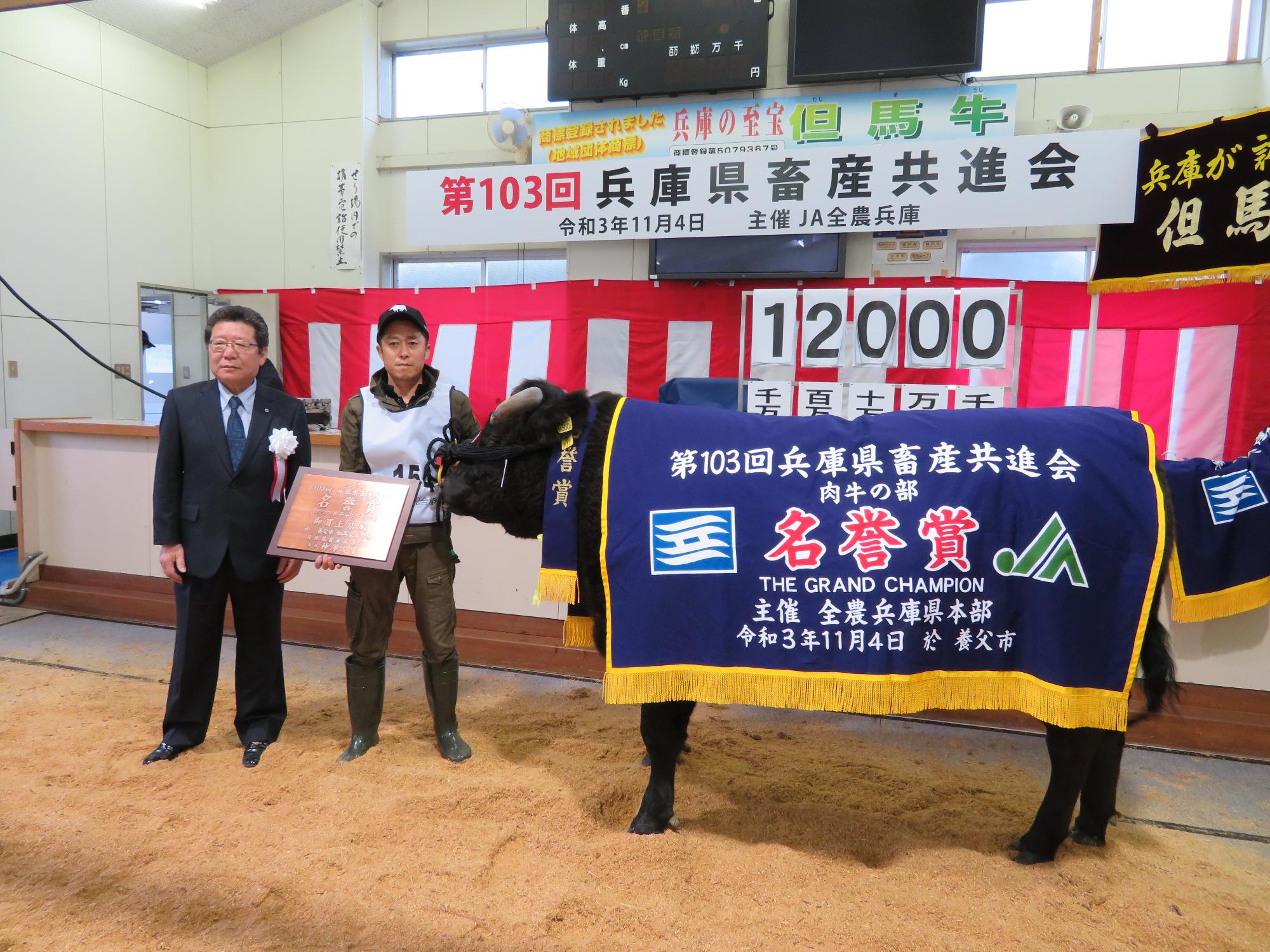 名誉賞と書かれた幕を背中からかけている牛の隣に、盾を持った畜産家の男性と、関係者の男性が立っている