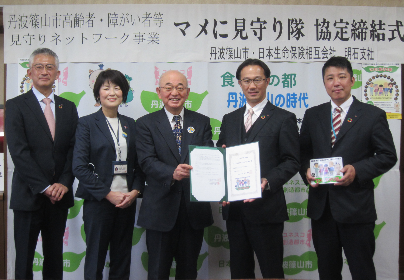 市長と事業所の社長が締結書を一緒に持っていて、そのまわりに社員の方が立っている。