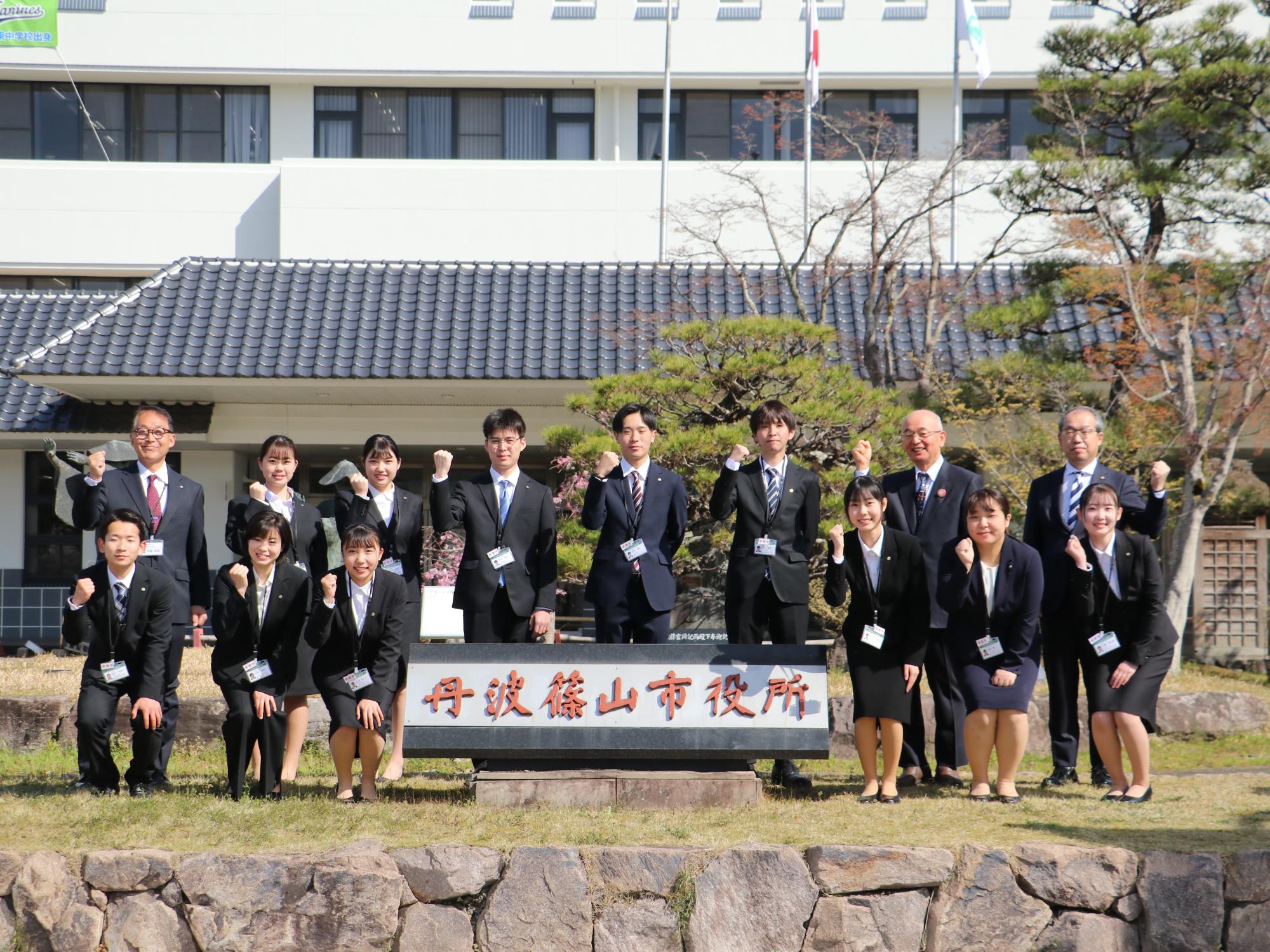 市役所の正面玄関前で三役と新入職員が記念撮影をしている