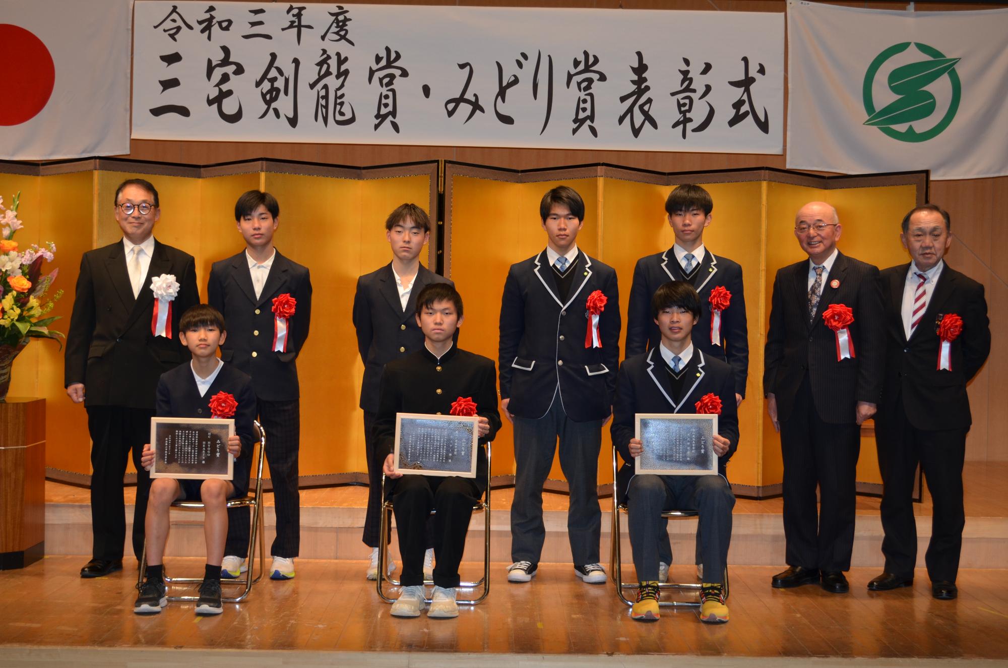 受賞者3名が表彰盾を座ってひざにかかげており、後列に関係者、市長、教育長、議長が立っている