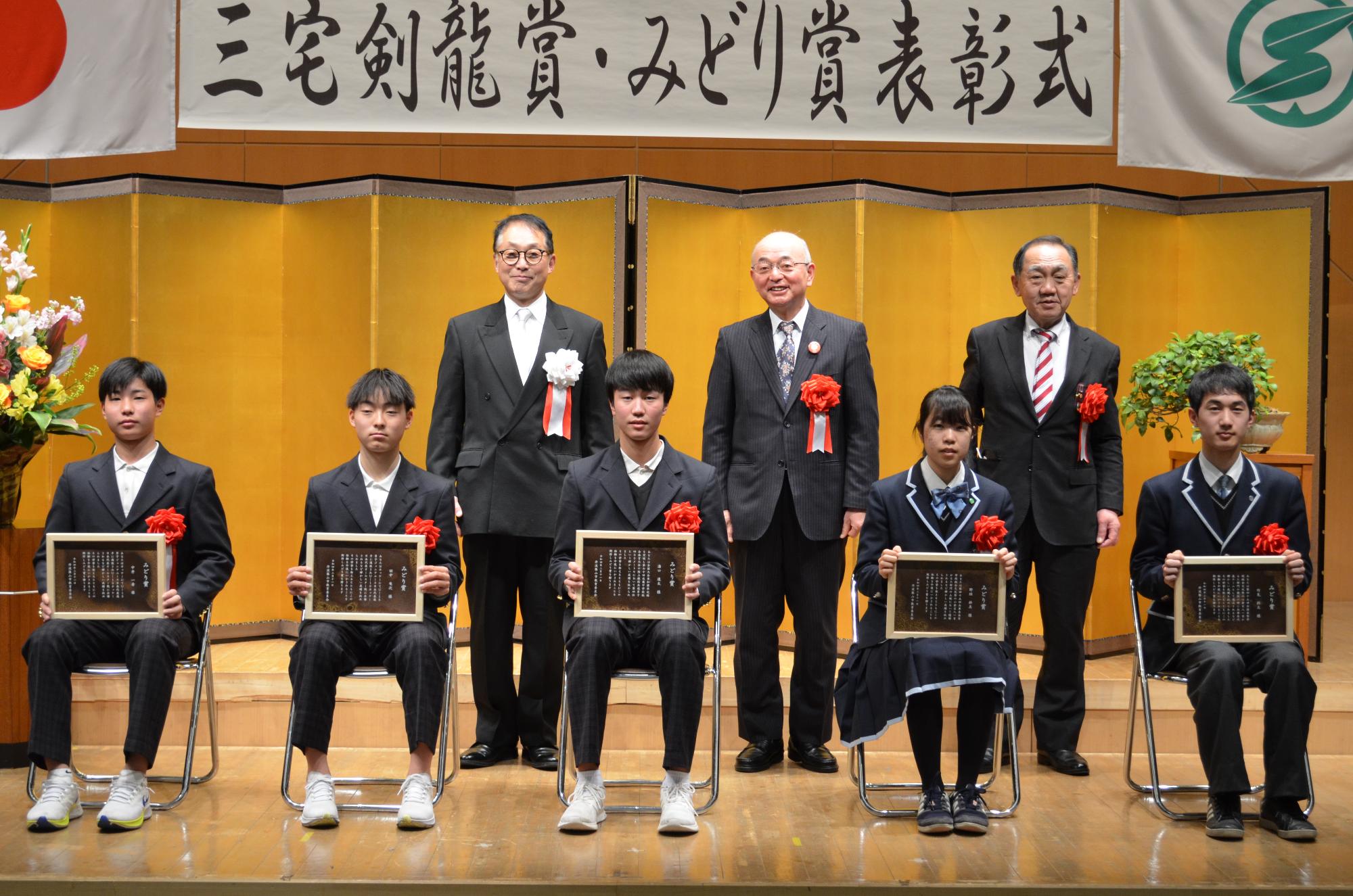 受賞者5名がいすに座って表彰盾をひざにかかげている。後列に市長、教育長、議長が並んでいる