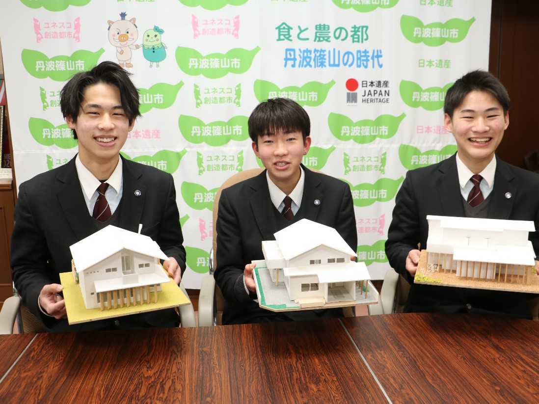 設計した家の模型を手に持った3人の男子高校生が椅子に座っている