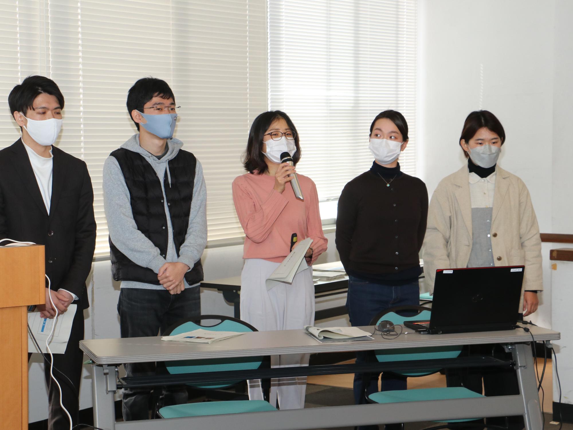 中央の女性がマイクを持って発表してい。まわりに4名立っている