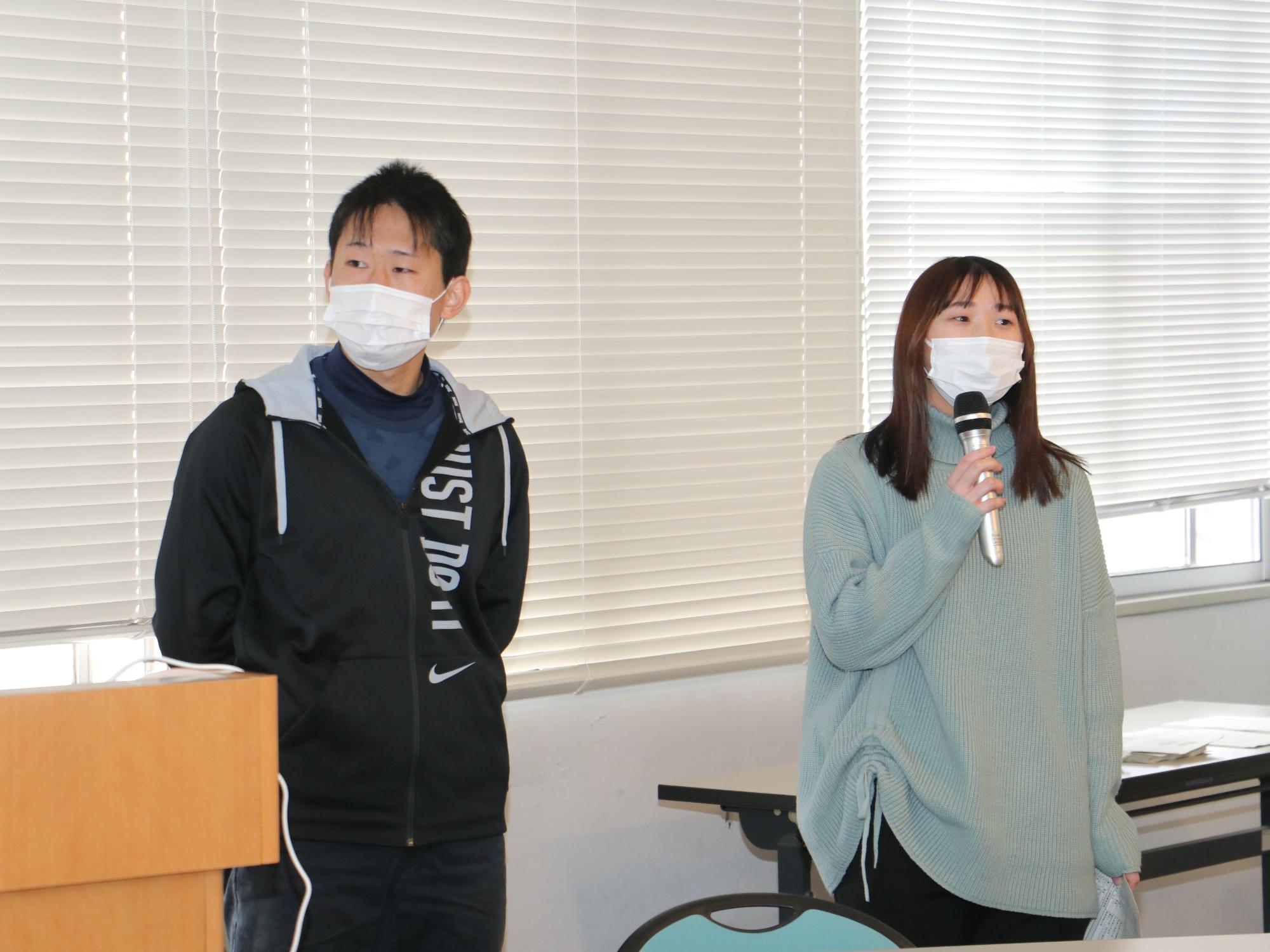 女性がマイクを持って発表している。隣に男性が立っている