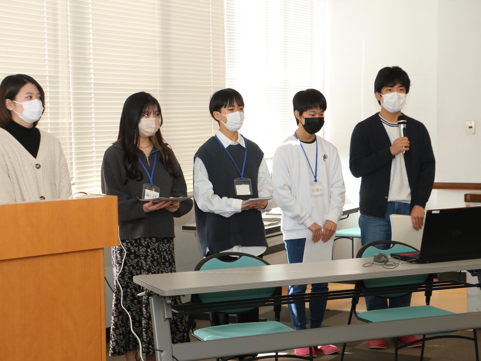 一人の男性がマイクを持って話していて、横に4名が立っている