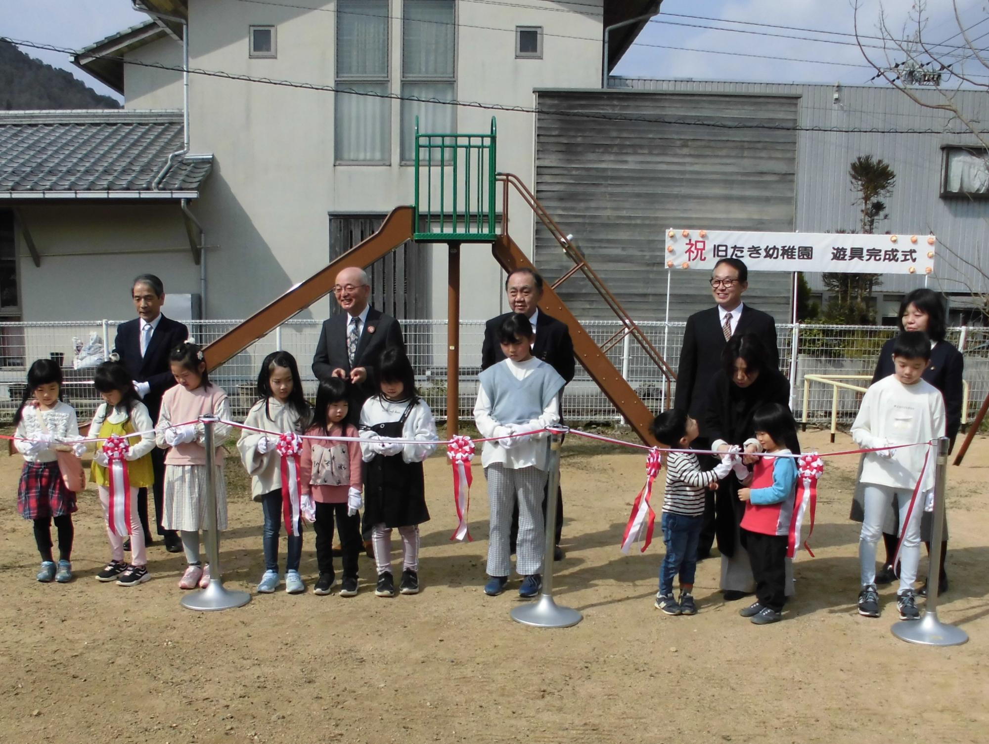 完成したすべり台の前で、子ども達がならんでテープカットをしようしている。後ろには関係者が並んでいる