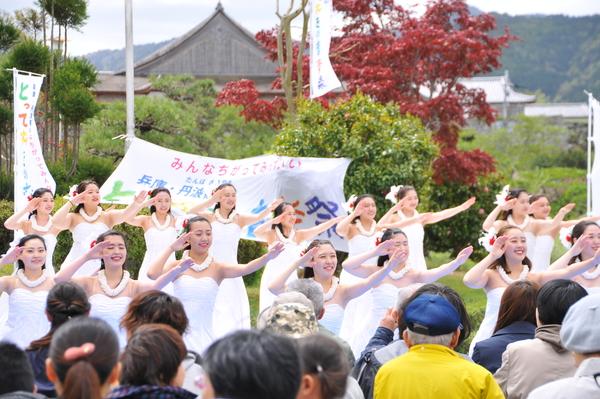 外のステージにて髪飾りをつけ白色の首飾りに白い衣装を着た女性らがフラダンスを踊っているのを見ている人々の写真