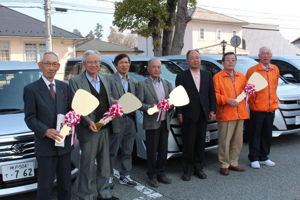 乗用車の前に7名の方が並んでおり、うち、4名の方が赤白テープのついた鍵を持ち、笑顔で写っている集合写真