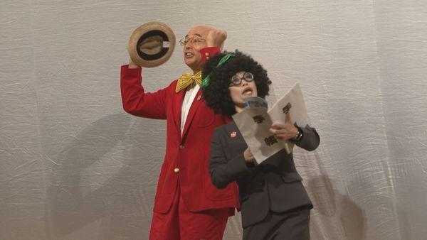 帽子を右手に持った蝶ネクタイに赤いブレザー姿の市長がガッツポーズをしており、手帳を持ったパーマヘアーにスーツ姿の秘書が声を上げている写真