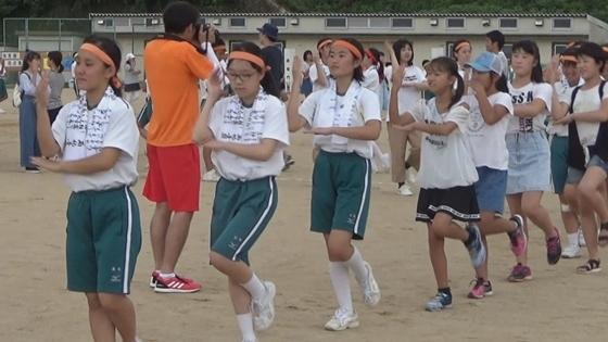 中学校の校庭に頭にハチマキをつけた体操服の中学生が西紀温度を円になり地域の方と踊っている映像写真