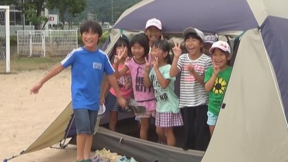 学校の校庭にテントを張り、その中から小学生がピースサインをしているところが写しだされているの映像写真