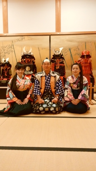 畳の上に鎧兜が4体置いてあり、その前で二名の袴姿の女性と市長が正座している写真
