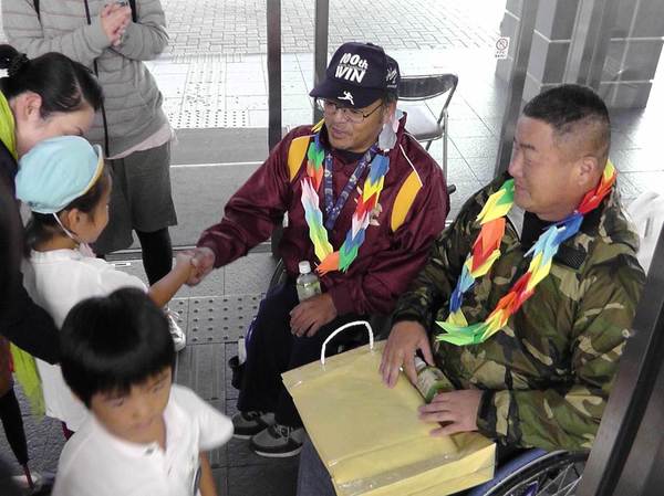 園児から色とりどりの折鶴のレイをもらっており、奥の帽子をかぶった男性が、水色の帽子の園児と握手をしている様子の写真