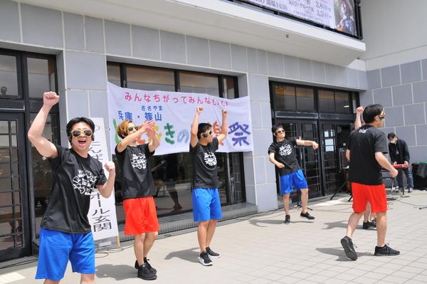 ホール玄関にてサングラスをかけ黒色のTシャツに赤や青の短パンをはいた男性5名が手を上げたり、拍手したりパフォーマンスしている写真