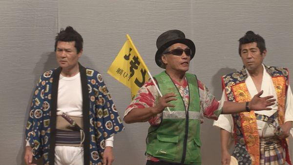 2人の侍の間にいる工事現場用の緑のベストにサングラスをかけ黒いハットを被った男性が交通安全の黄色い旗を持って話している写真