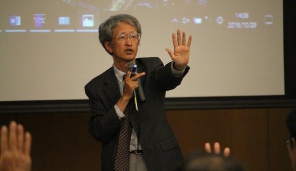 東京大学の堀 繁教授がマイクを右に持ち、左手を広げ手の平を見せ講演されている写真