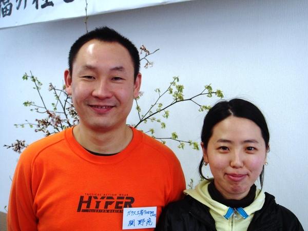 左側にオレンジ色の洋服を着ている関野 亮さんと右側に黄色いパーカーを着ている奥様が笑顔で写っている写真 拡大画像