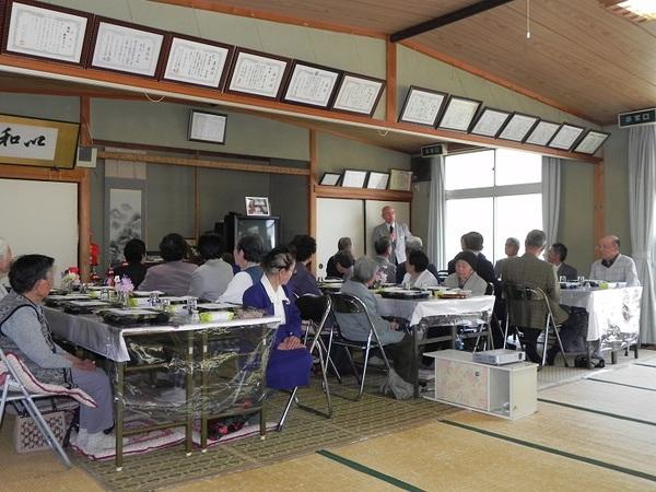 お部屋にはたくさんの賞状が飾られ、年配の方々が代表で話している男性のお話しを聞いている写真