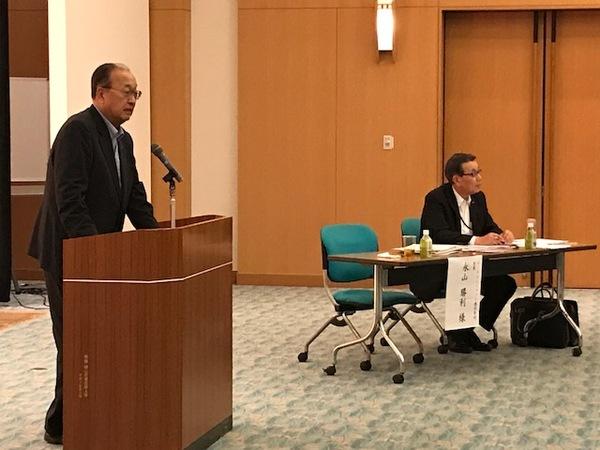 ルートインジャパン株式会社の永山勝利会長が立って話をしていてる横に男性が座っている写真