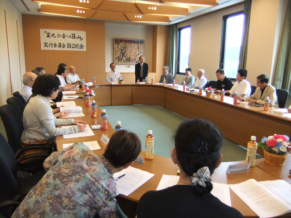 市民センターにて男性が立って話をされていて、テーブルに資料とお茶が置いてあり参加された方が座って話を聞いている写真