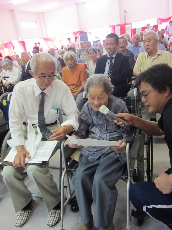 沢山のご高齢者のまえで、紙を見て読み上げる、車椅子の高齢の女性の写真