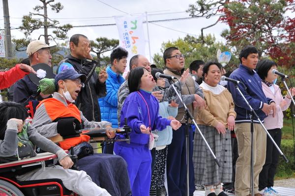外のステージにスタンドマイクが3台あり、車いすに乗った男性らたくさんの人々が歌を歌っている写真