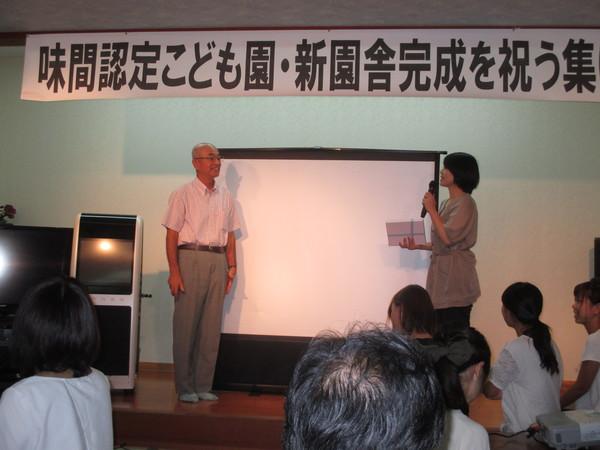 味間認定こども園・新園舎完成を祝う集と書かれた幕のかかった舞台の上でマイクを持った女性と市長が立っている写真