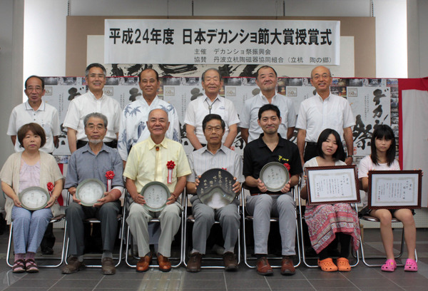 胸章リボンをつけ、賞状やまるいプレートをもって座る受賞者と関係者の集合写真