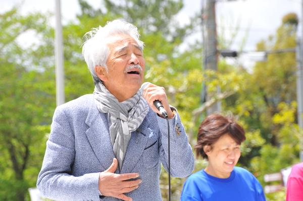 外のステージでスーツにストールを巻いた男性がマイクを持って歌を熱唱しているのを人々が聞いている写真