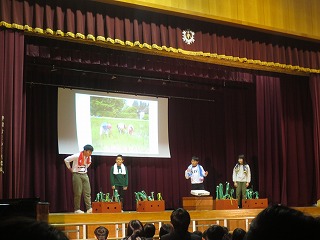 首からタオルをさげた4人の5年生の子どもたちが、農業をしている写真をスクリーンに映して、劇をしている写真