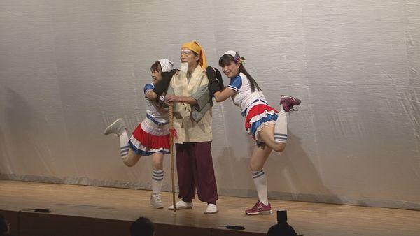 舞台に杖を突いて立っている黄門さまを真ん中にチアガール風の衣装を着て日本タオルを頭にのせた女性が黄門さまに寄り添って片足を上げて立っている写真