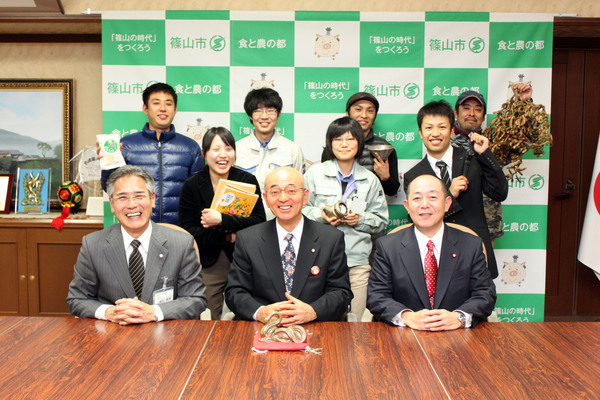 テーブルに着席している酒井市長、足立市議会議長、前川教育長とその後ろに立っている帰路または転入された7人との記念撮影している写真