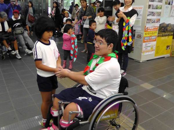 車いすの朽木くんが表彰を受けて折り鶴のレイを首にかけており、女の子の園児と握手をしている写真