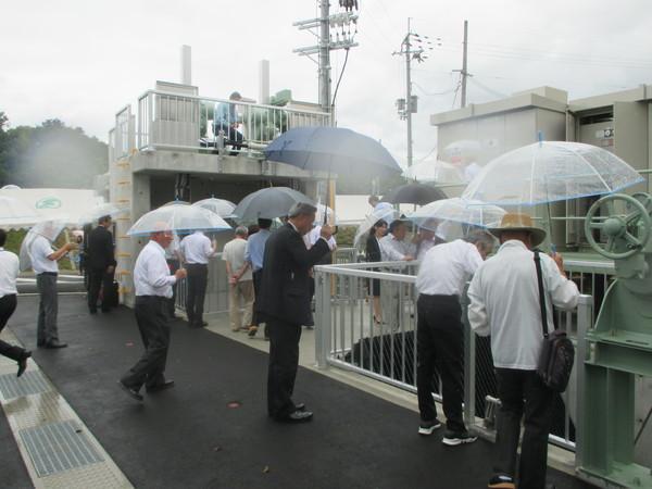 雨の中、傘を差した方々が排水ポンプ設置の機械を見ている写真