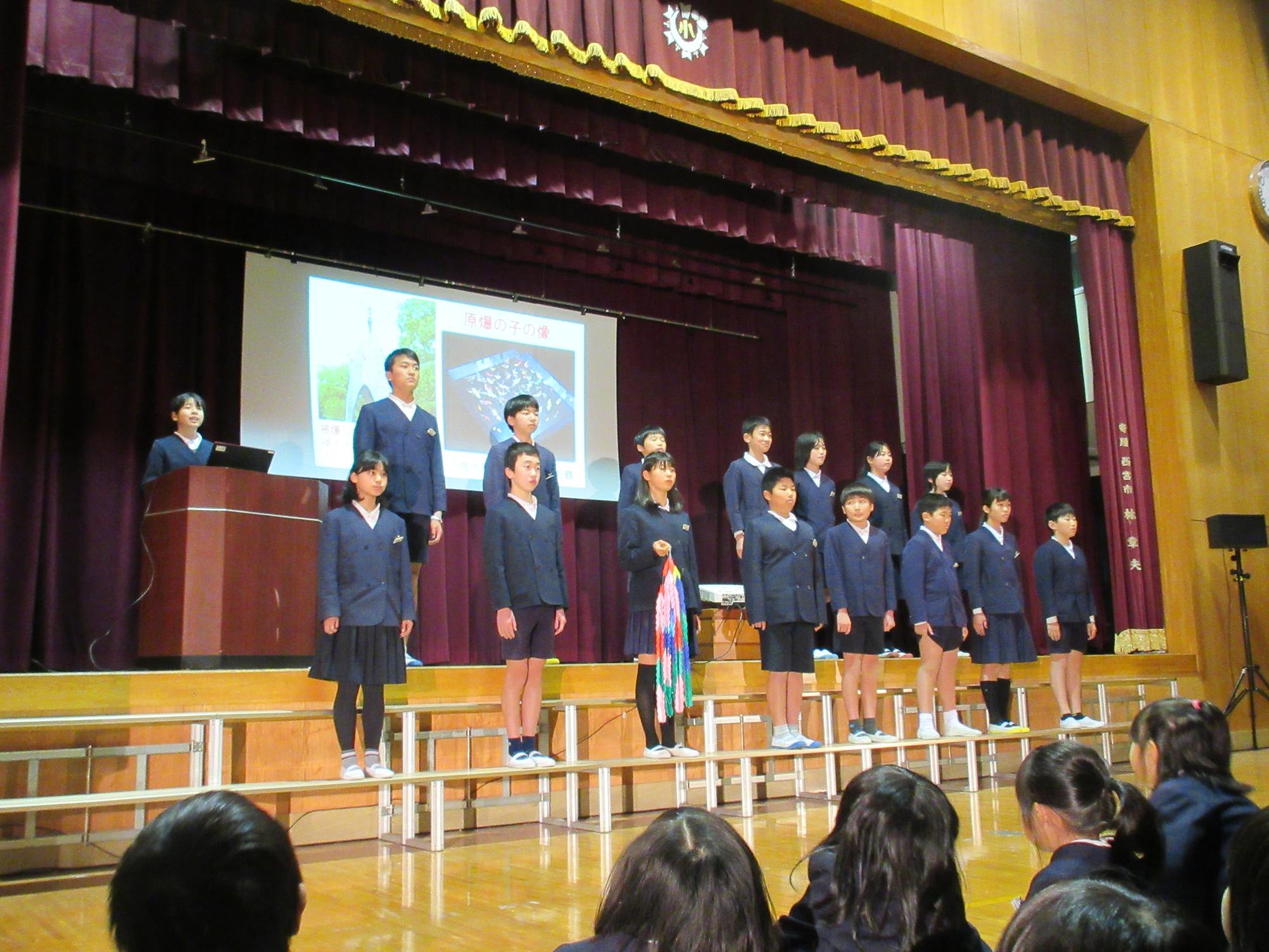 17人の6年生の子どもたちが、横2列に並んでおり、下の段の左から3番目の子が千羽鶴を手に持っている写真