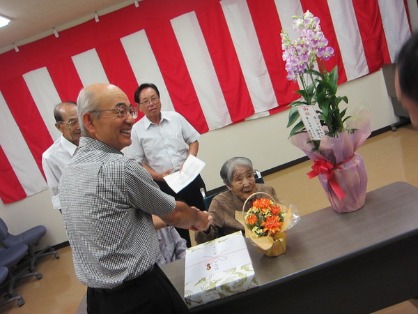 車いすに座る酒井 志づ子さんと握手をしている市長、机の上に置かれたお花と記念品の写真