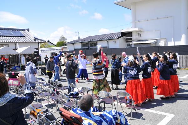 駐車場で拳を上げて踊っている若者を椅子に座って見ている人々の写真