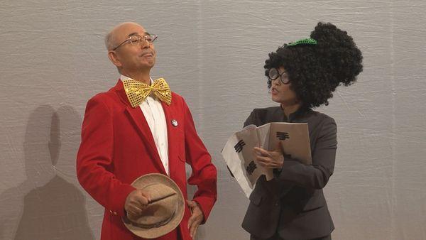 帽子を右手に持った蝶ネクタイに赤いブレザー姿の市長が手帳を持ったパーマヘアーにスーツ姿の秘書にスケジュールの確認をされている写真