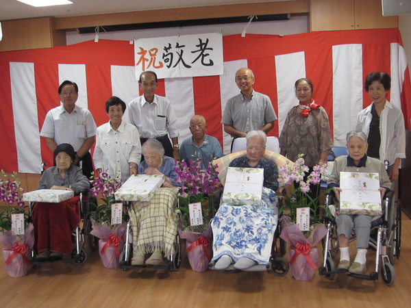 車いすの4名の高齢者が、送られた花の横で記念品を膝の上に置いて、後ろに立つ関係者や市町の写真