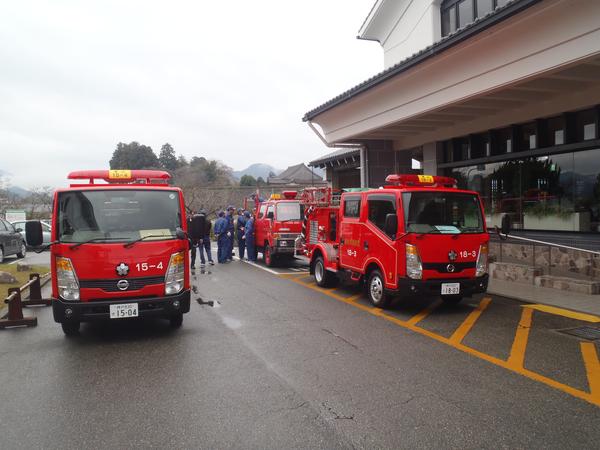 建物の前に駐車されている消防車3台と遠くに作業服姿の団員らが写っている写真