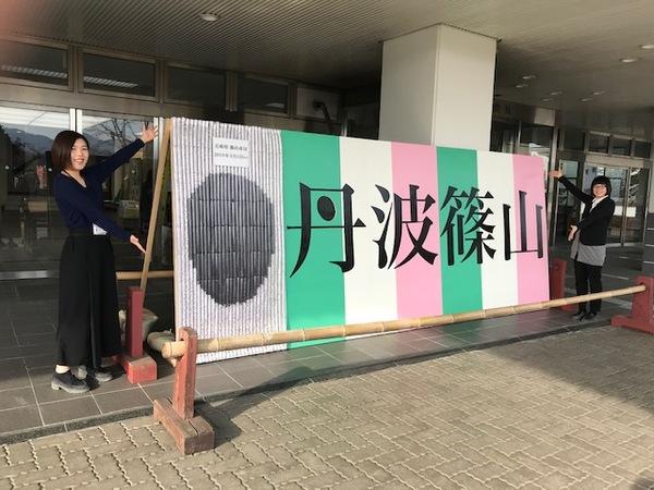 市役所本庁舎前に丹波篠山パネルが飾られ、両端で女性職員がポーズを取っている写真