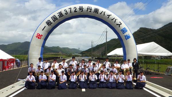 祝 一般国道372号 丹南バイパス開通と書かれたバルーントンネルの前に丹南中学校吹奏楽部の子供たちが楽器を持ち、ひざまずいている集合写真
